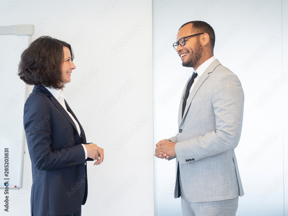 Two friendly confident professionals discussing business issues in conference room. Business man and woman standing indoors near white board and chatting. Business communication concept Two friendly confident professionals discussing business issues in conference room. Business man and woman standing indoors near white board and chatting. Business communication concept