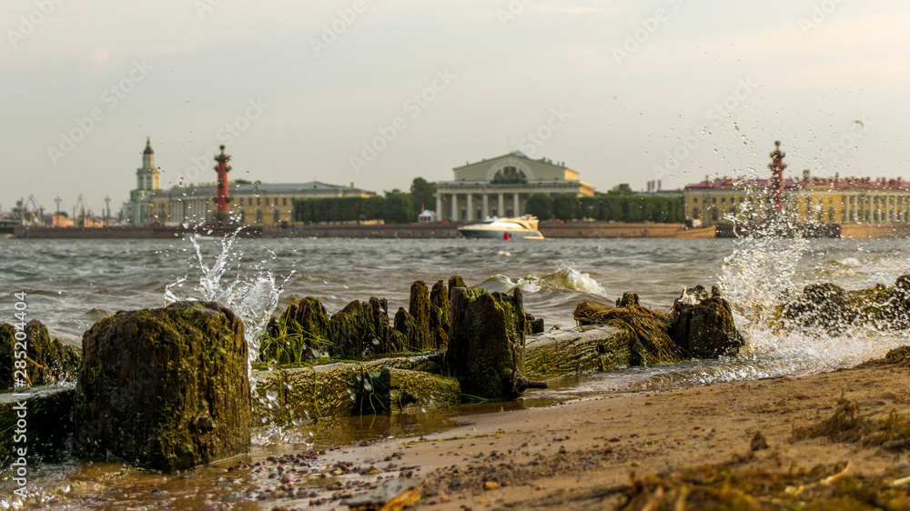 Fototapeta premium Saint Petersburg, Russia. View of the Spit of Vasilyevsky Island. The beach of the Peter and Paul Fortress. Neva River. Splashing waves breaking on the shore. Boat sailing on the Neva River.