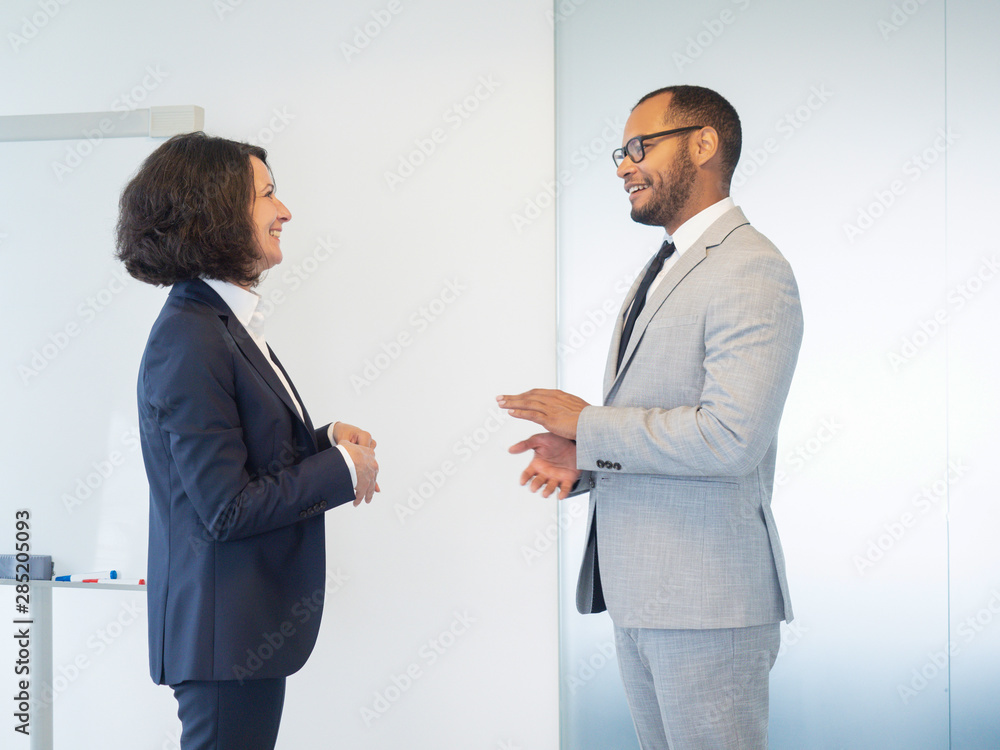 Two friendly business colleagues discussing training in conference room. Business man and woman standing indoors near white board, talking and laughing. Business colleagues concept Two friendly business colleagues discussing training in conference room. Business man and woman standing indoors near white board, talking and laughing. Business colleagues concept