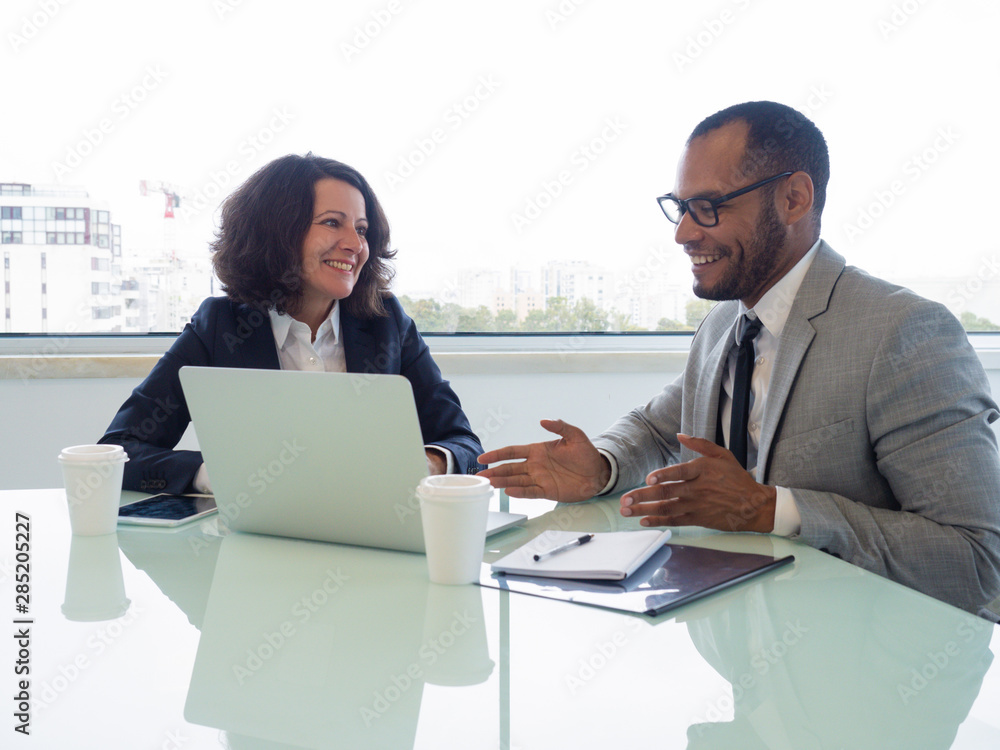 Happy coworkers discussing new business software. Business man and woman sitting at meeting table with open laptop, talking and smiling. Business software concept Happy coworkers discussing new business software. Business man and woman sitting at meeting table with open laptop, talking and smiling. Business software concept