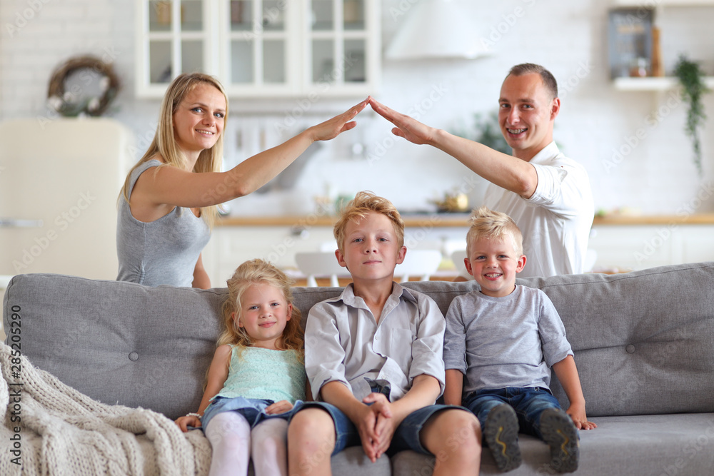 Happy full family with three kids sitting on sofa, mom and dad making roof figure with hands arms over heads. New building residential house purchase apartment and housing concept. Happy full family with three kids sitting on sofa, mom and dad making roof figure with hands arms over heads. New building residential house purchase apartment and housing concept.