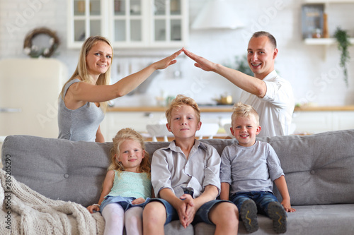 Happy full family with three kids sitting on sofa, mom and dad making roof figure with hands arms over heads. New building residential house purchase apartment and housing concept.