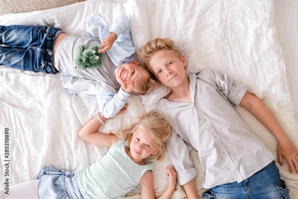 Three cheerful blonde kids are lying together on bed, looking at camera and smiling. Two boys and girl. Happy family, brothers and sister Three cheerful blonde kids are lying together on bed, looking at camera and smiling. Two boys and girl. Happy family, brothers and sister