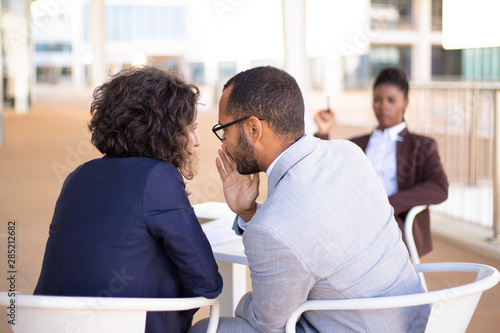 Employees gossiping about young female colleague. Business man and woman whispering, African American employee sitting in background. Office rumors concept photo