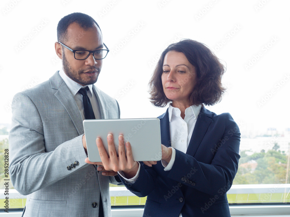 Business team watching presentation on tablet and discussing project. Business man and woman standing indoors, looking at tablet screen and talking. Communication and teamwork concept Business team watching presentation on tablet and discussing project. Business man and woman standing indoors, looking at tablet screen and talking. Communication and teamwork concept