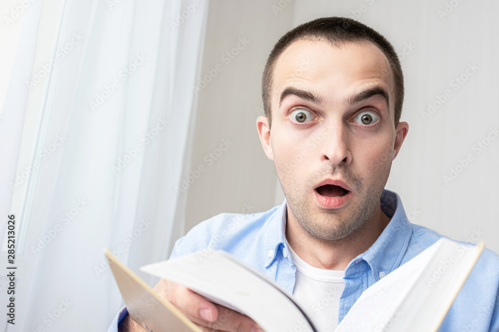 Shocked stunned student because of a very thick book, the student does not like to read and learn, looking at the camera. The concept of heavy loads in education, toned Shocked stunned student because of a very thick book, the student does not like to read and learn, looking at the camera. The concept of heavy loads in education, toned