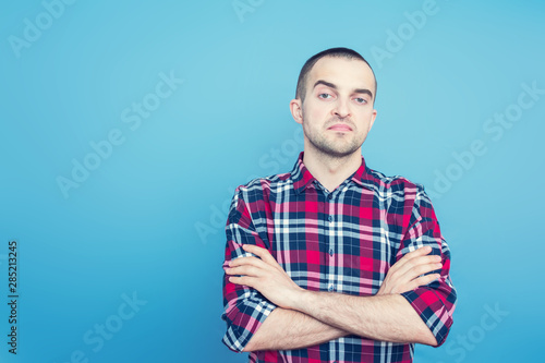 Attractive guy in plaid shirt haughtily looks at the camera, standing and having his arms crossed, front view, blue background, copy space, for advertising, for slogan, toned