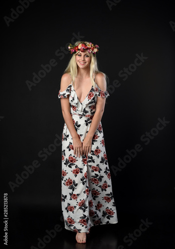 full length portrait of a blonde girl wearing a long white and floral dress, wearing a flower crown. Standing pose on a black studio background.