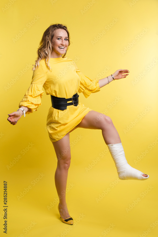 Full length portrait of stunning young woman in yellow dress with one leg in plaster. She is posing with bent broken leg in plaster showing positive attitude. Cheerful lady in dress with plaster