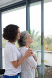 Pensive mother and daughter looking at window. Side view of thoughtful senior mother and adult daughter standing together and looking at window. Togetherness concept