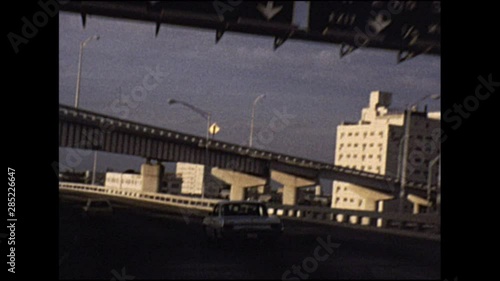 Driving along highway following traffic under urban bridge in 1970s USA, vintage footage.