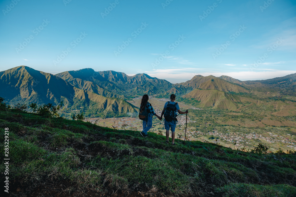 Obraz premium Young couple with backpacks hiking in the mountains and enjoying valley view. Back view of young tourist couple with backpacks, athletic man and slim girl stand holding hands on rocky mountai