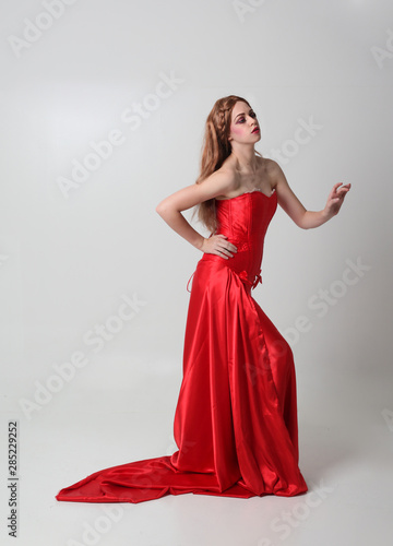 full length portrait of a girl wearing a long red silk gown, Standing pose on a grey studio background.