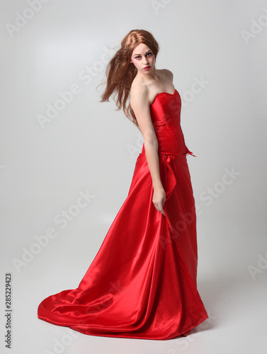 full length portrait of a girl wearing a long red silk gown, Standing pose on a grey studio background.