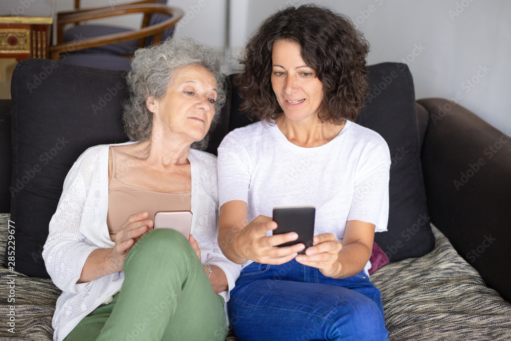 Smiling mother and daughter using smartphones. Cheerful senior and middle aged women sitting together on couch and using mobile phones. Technology concept Smiling mother and daughter using smartphones. Cheerful senior and middle aged women sitting together on couch and using mobile phones. Technology concept