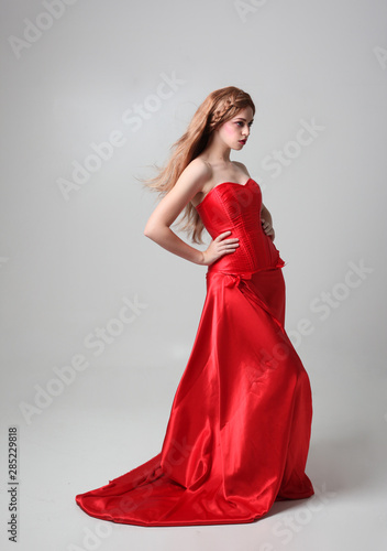 full length portrait of a  girl wearing a long red silk gown, Standing pose on a grey studio background. © faestock