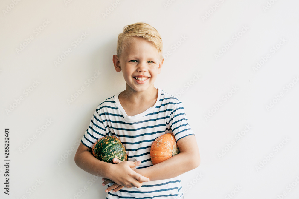 Caucasian blond boy against the background of a white wall holds two pumpkins under his armpits and laughs. Lifestyle portrait of a child for autumn harvest. Copy space for text. Caucasian blond boy against the background of a white wall holds two pumpkins under his armpits and laughs. Lifestyle portrait of a child for autumn harvest. Copy space for text.
