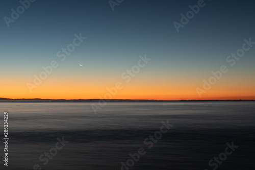 A long exposure captures the crescent moon overlooking over the beautiful gradient created by the sun after it has set into the horizon on North Stradbroke Island  Queensland  Australia