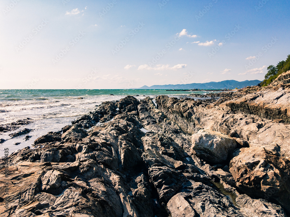 A rocky, impassable shore with green plants against the backdrop of a beautiful turquoise sea with little excitement in cloudy weather on a sunny summer day.