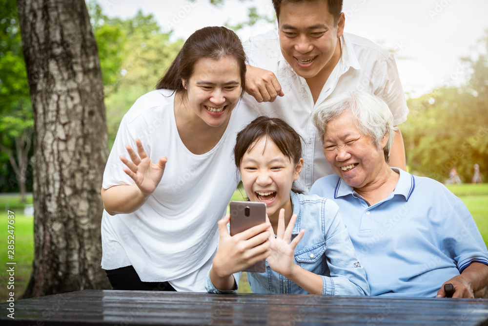 Fototapeta premium Communication technology,happy asian family laughing with smartphone,daughter,father,mother,grandmother talking to each other through a video call on a smartphone,parents with child girl enjoying park