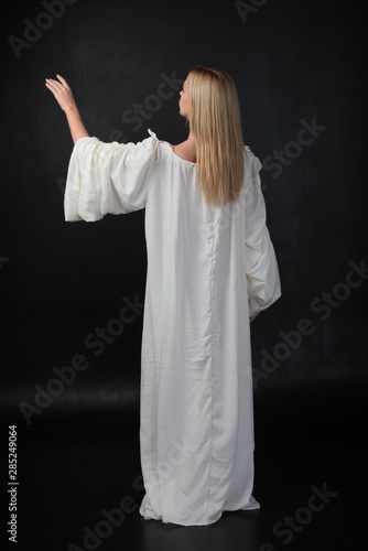 full length portrait of blonde girl wearing long white flowing robe. standing pose against a black studio background. photo