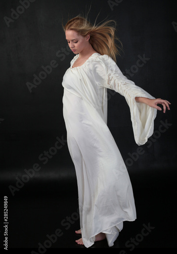 full length portrait of blonde girl wearing long white flowing robe. standing pose against a black studio background.