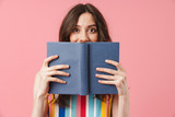 Beautiful happy emotional young cute woman posing isolated over pink wall background covering face with book.