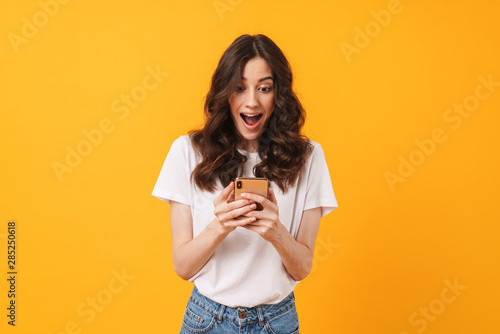 Happy shocked emotional young woman posing isolated over yellow wall background using mobile phone.