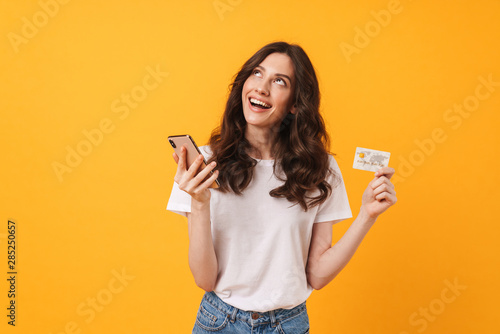 Dreaming young woman posing isolated over yellow wall background using mobile phone holding debit card.