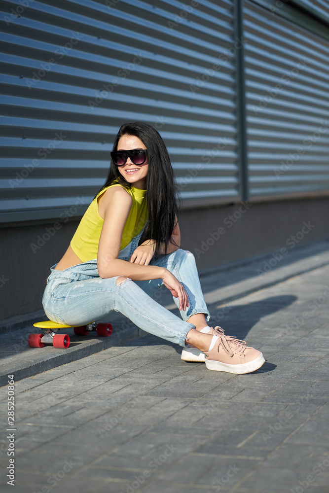 Fototapeta premium Beautiful stylish smilling brunette girl with long hair in trendy sunglasses wearing yellow top, blue denim overalls and fashionable pink sneakers sitting on yellow skateboard with red wheels.