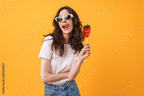 Happy cute young woman posing isolated over yellow wall background holding candy lollipop.