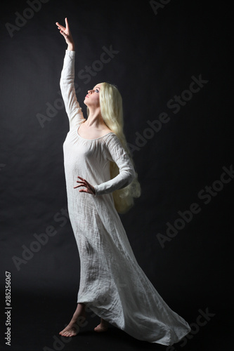 Ghostly full length portrait of a woman with long blonde hair wearing a white robe. Standing pose against a black studio background. 