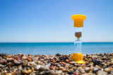 yellow hourglass with crumbling sand standing on the wet sea pebbles with the sea and the clear sky in the background