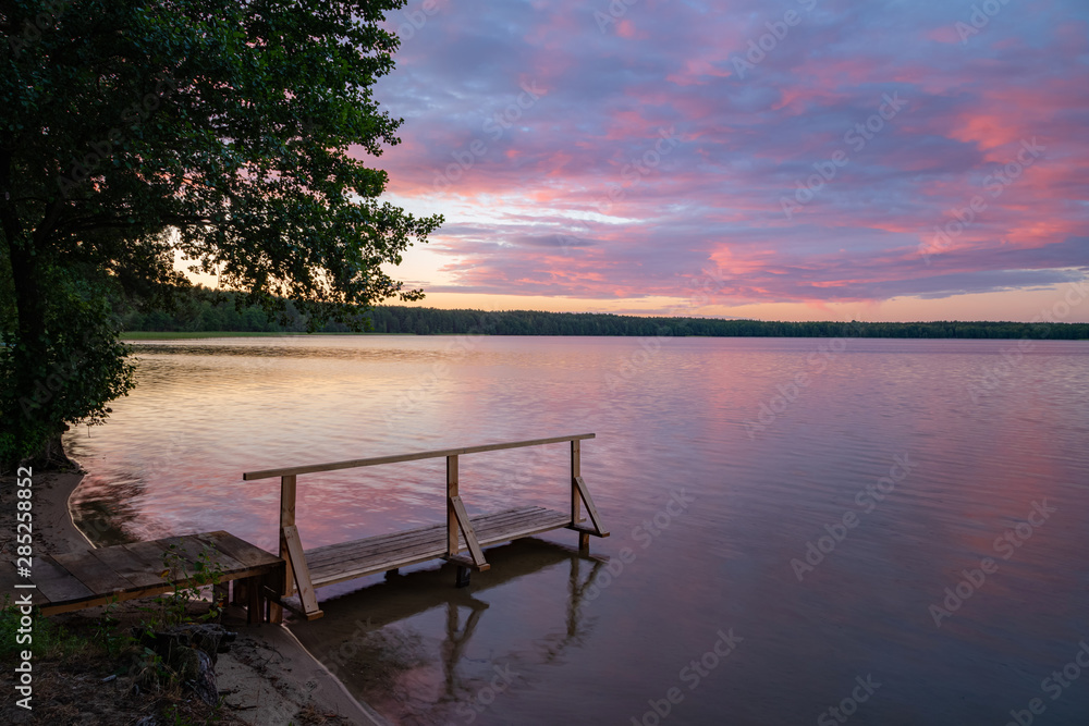 Wooden pier by the lake with burning sunrise sky and forest in the background. Colourful sunset sky by the water shore. Peaceful time camping in nature