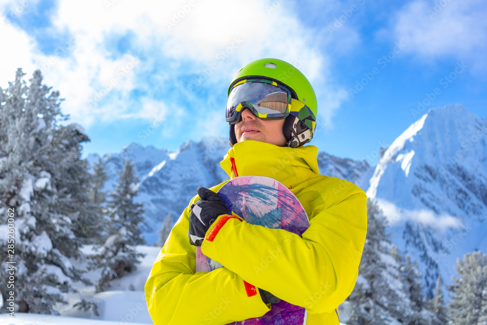 Snowboarder man with ski glasses in green helmet make photoshoot on top in Alps mountains. On the background of mountains. Close up view.