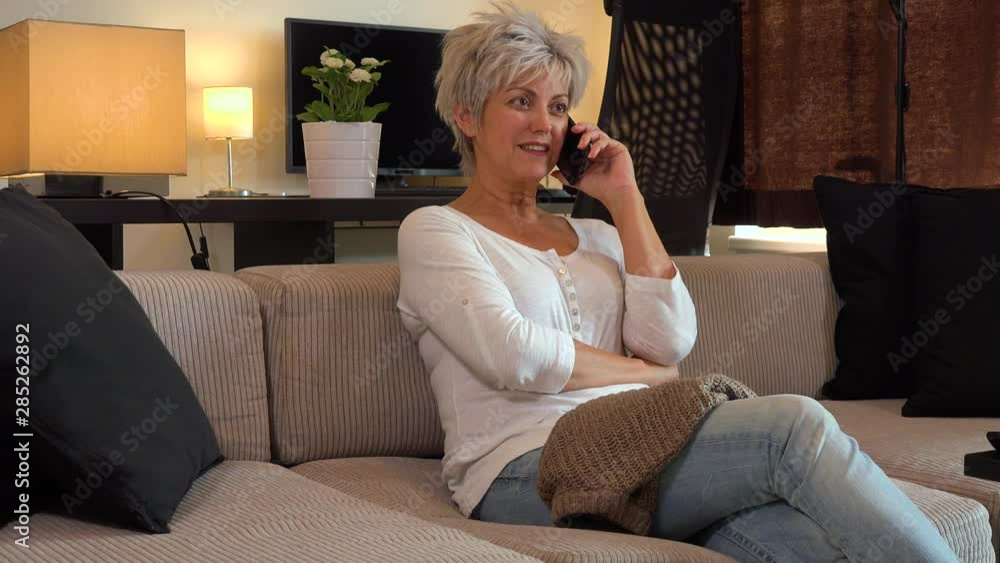 A middle-aged woman sits on a couch in an apartment living room and talks on a smartphone with a smile A middle-aged woman sits on a couch in an apartment living room and talks on a smartphone with a smile
