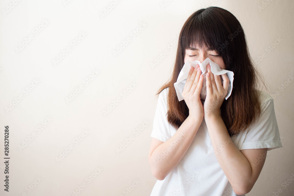 Young Asian woman sneezing and blowing her nose w/ tissue paper. Cause of runny nose include common cold, flu, allergies, sinusitis, asthma, air pollution or influenza. Health care. Copy space. Young Asian woman sneezing and blowing her nose w/ tissue paper. Cause of runny nose include common cold, flu, allergies, sinusitis, asthma, air pollution or influenza. Health care. Copy space.
