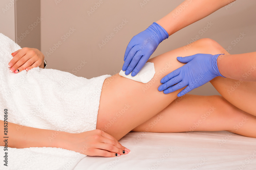 Fototapeta premium Beautician waxing female legs in spa center. Removing unnecessary hair on the legs. Procedure sugaring in a beauty salon. Sugar depilation. Depilatory sugar paste. Epilation with liquate sugar at legs