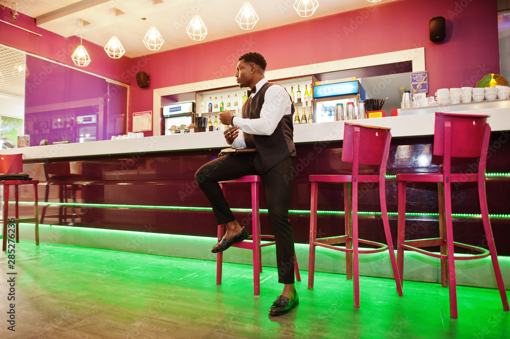Fototapeta premium Young handsome african man wearing white shirt, black vest and bow tie with wallet purse posed against bar counter at night club.