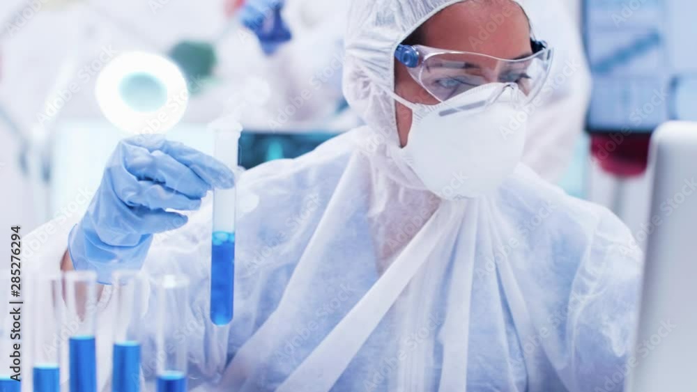Female researcher in cover all equipment holding a test tube with smoking blue solution. Scientist typing on computer. Female researcher in cover all equipment holding a test tube with smoking blue solution. Scientist typing on computer.