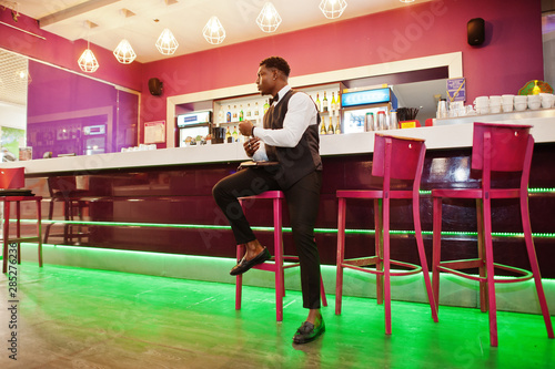 Young handsome african man wearing white shirt, black vest and bow tie with wallet purse posed against bar counter at night club. © AS Photo Family