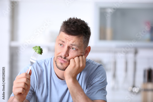Portrait of unhappy man looking at broccoli on fork in kitchen