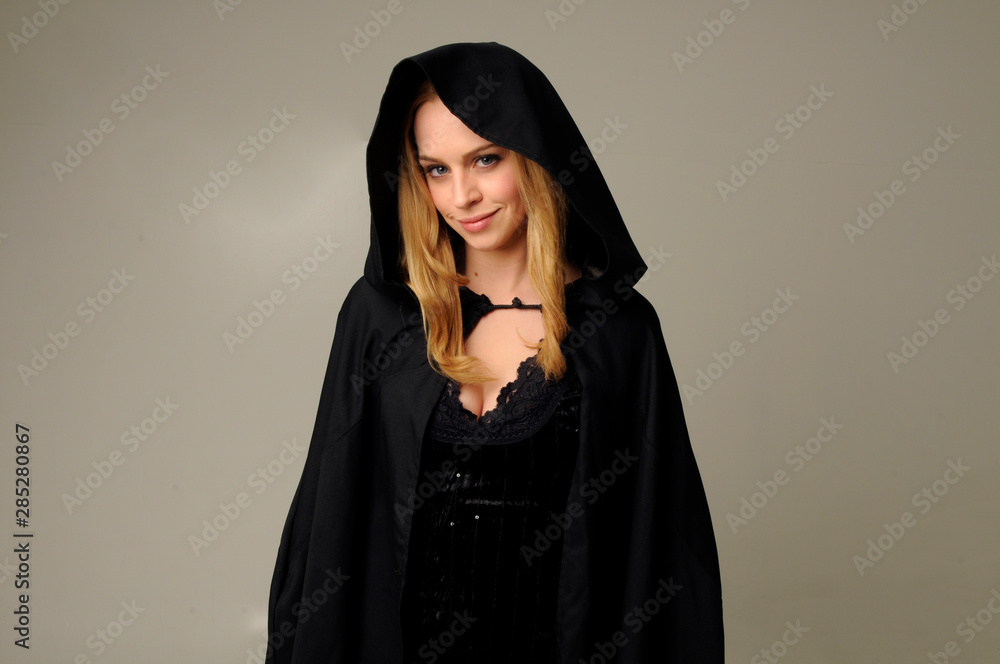portrait of blonde woman wearing black hood, halloween witch costume. creamy studio background. portrait of blonde woman wearing black hood, halloween witch costume. creamy studio background.