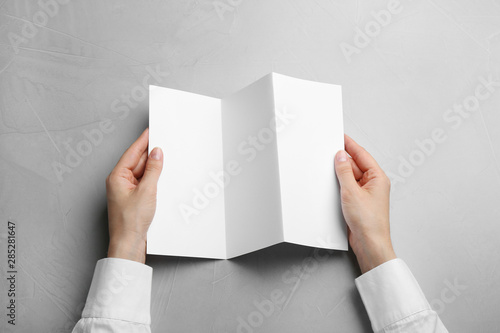 Young woman holding blank brochure at grey table, top view. Mock up for design