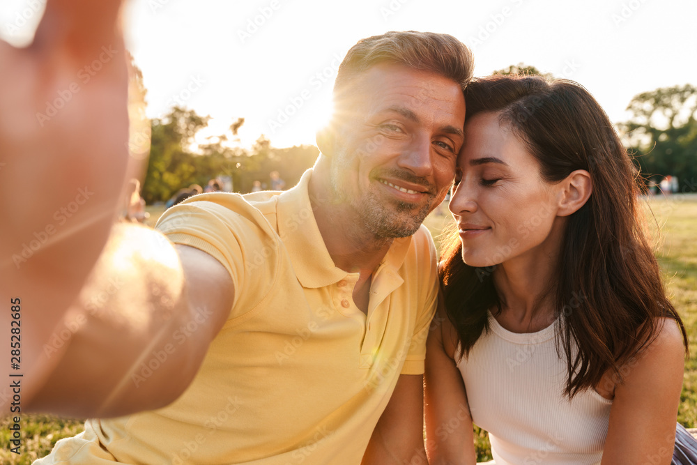 Smiling cute adult loving couple outdoors in a beautiful green nature park take a selfie by camera. - obrazy, fototapety, plakaty 