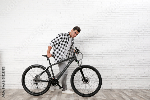 Handsome young man with modern bicycle near white brick wall indoors