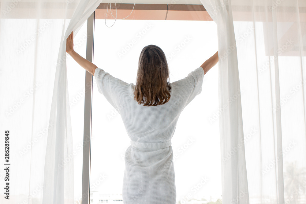 Woman open white curtains at the window, the morning after waking up.
