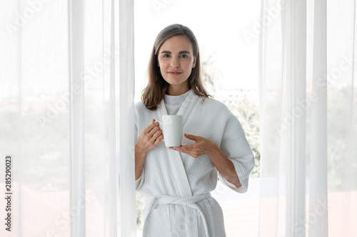 Caucasian woman in pajamas holding a glass of white coffee ready to drink by the window.