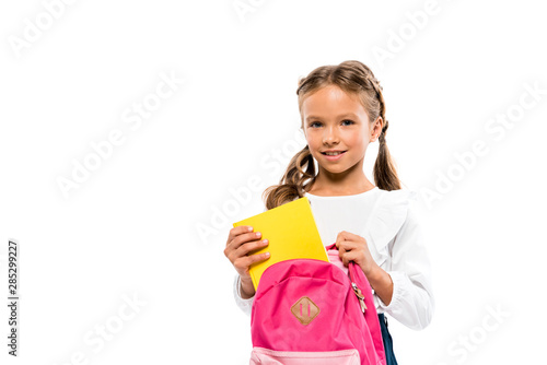 Bilde på lerret cheerful child putting book in pink backpack while standing isolated on white