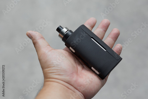 high end black regulated  box mods with rebuildable dripping atomizer in hand on light grey texture background, vaping device, selective focus photo